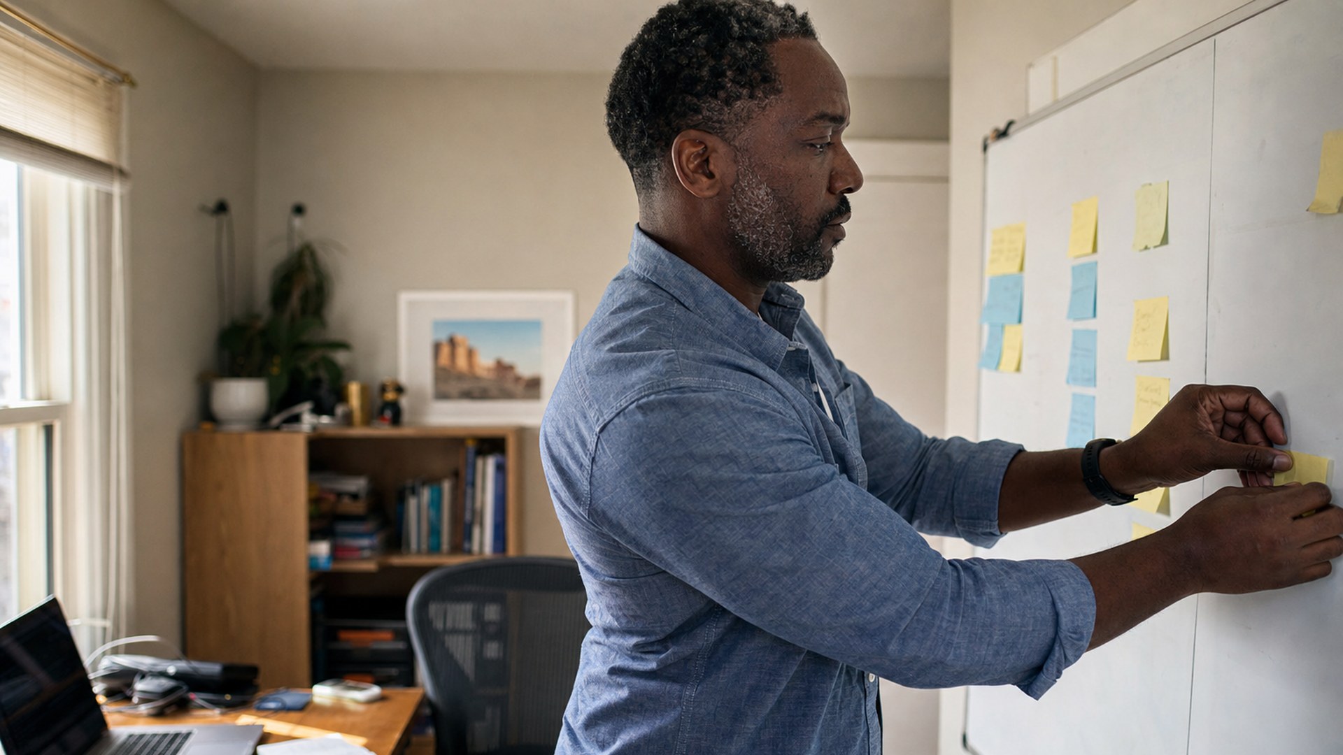 A freelance developer adding a sticky note to a whiteboard of project tasks in his home office